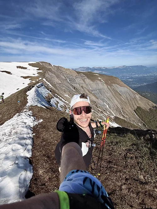 Louis Gronemberg, souriant avec équipement de trail en montagne, prise de selfie avec paysage de montagnes enneigées et ciel bleu derrière lui, lors de la SkyRace de Matheysins.