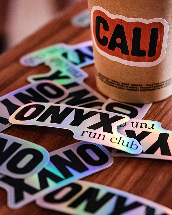 Une tasse en carton avec une étiquette ‘CALI’ posée sur une surface en bois, entourée de plusieurs autocollants avec des mots comme ‘ONYX’, ‘run’, ‘club’.