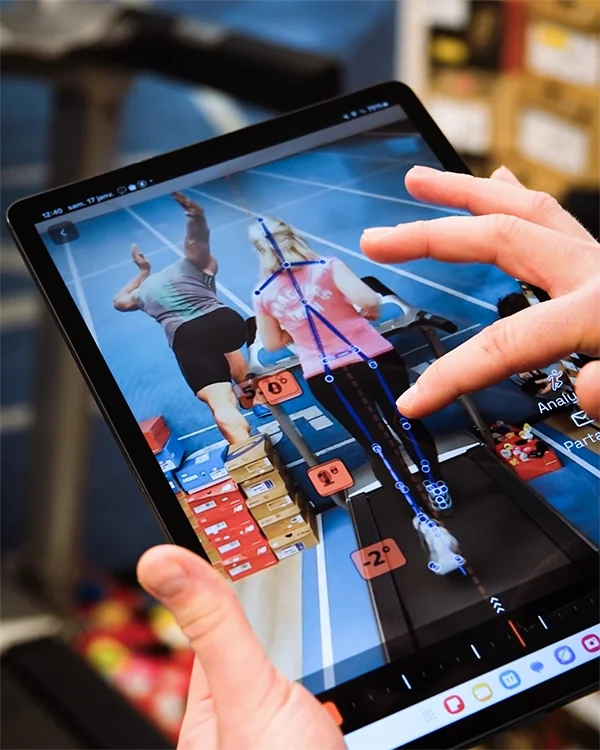 Une personne utilise une tablette pour analyser un saut en longueur lors d'une compétition sportive en intérieur, avec une piste bleue, des boîtes empilées et des athlètes en action, lors d'une analyse de foulée avec Louis Gronemberger chez UTS.