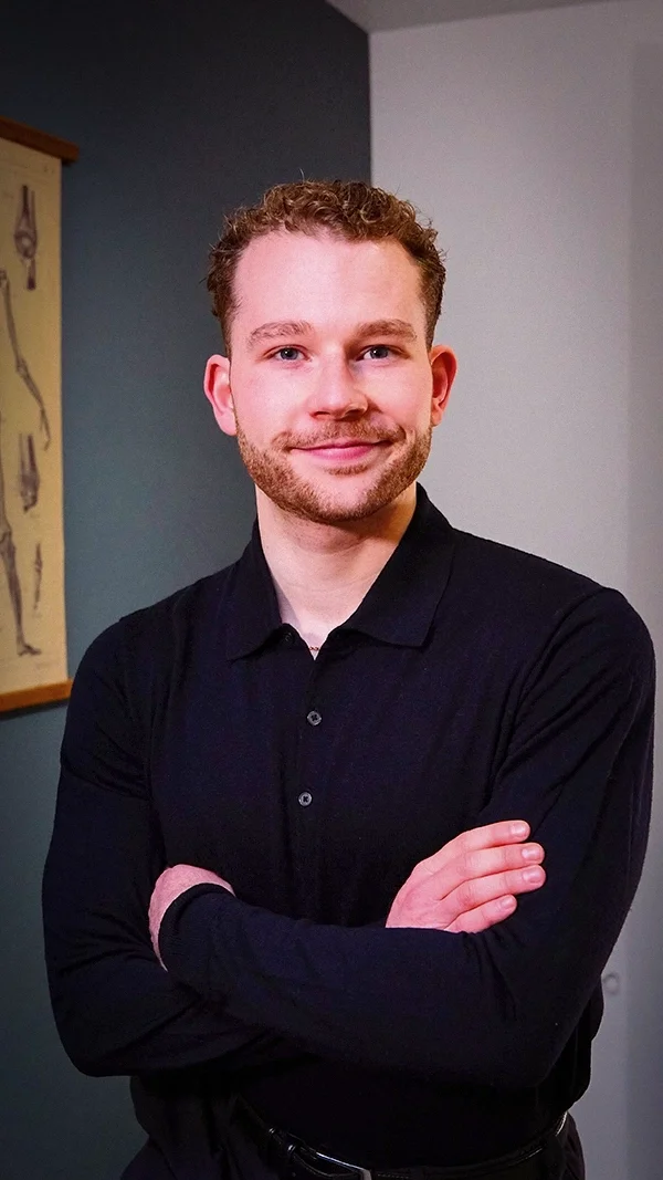 Louis Gronemberger, jeune homme souriant avec des cheveux bouclés, portant une chemise noire, posé avec les bras croisés devant un mur gris à l'intérieur.