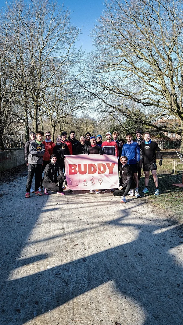 Groupe de personnes en tenue de course, tenant une bannière rouge et blanche avec le texte « BUDDY », posant pour une photo en plein air avec des arbres sans feuilles et un ciel bleu en arrière-plan, lors d'un run avec Onyx à Stockel.