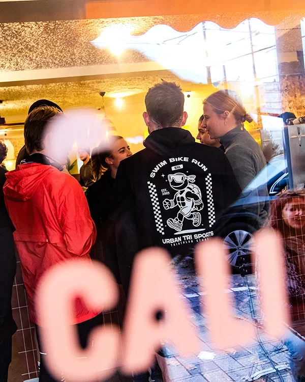Groupe de personnes discutant à l'intérieur d'un café ou restaurant, vue à travers une vitre avec une inscription "Cali" en flou, à Stockel avec le run club Onyx.