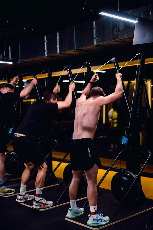 Trois hommes s'entraînent sur des machines d'exercice dans une salle de sport, en particulier des rameurs, à l'Hyrox Lab du Mix Brussels.