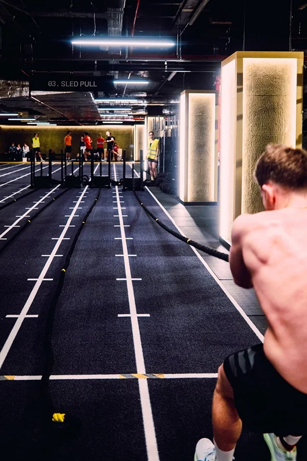 Salle de sport, l'Hyrox Lab au Mix Brussels, avec un jeune homme qui tirent des poids, plusieurs personnes en arrière-plan, quelques murs en couleur et un éclairage moderne.