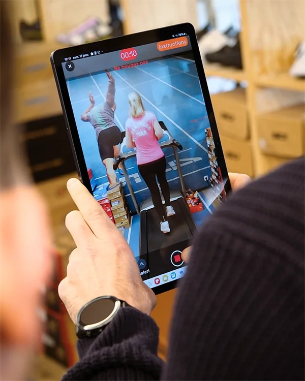 Une personne tient une tablette enregistrant une vidéo d'une autre personne marchant sur un tapis roulant, dans une salle avec des étagères en bois, lors d'une analyse de foulée avec le kiné Louis Gronemberger.