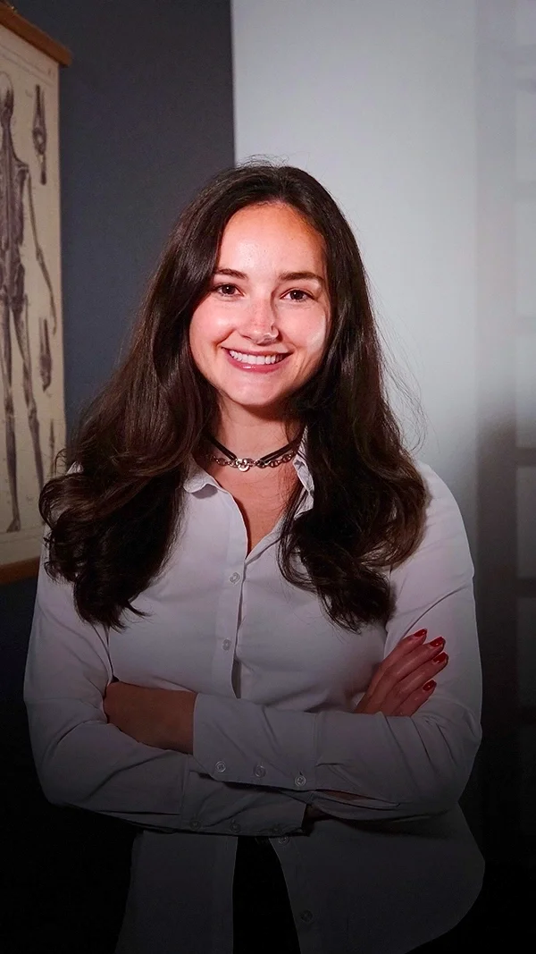 Marine Platiau, jeune femme avec des cheveux longs et bruns, portant une chemise blanche, souriant, les bras croisés, dans une pièce avec un décor artistique au mur.