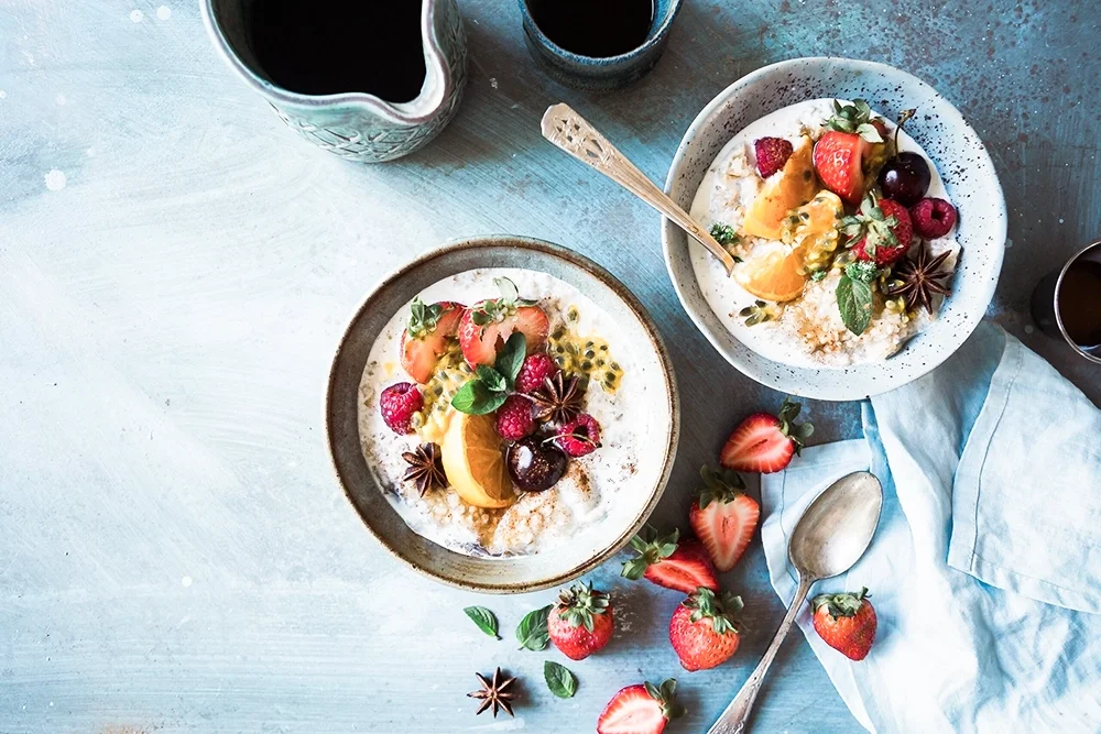 Bol de fruits frais et céréales, avec fraises, oranges et autres fruits, sur une table avec des cuillères, deux verres de boisson, et quelques fruits autour.