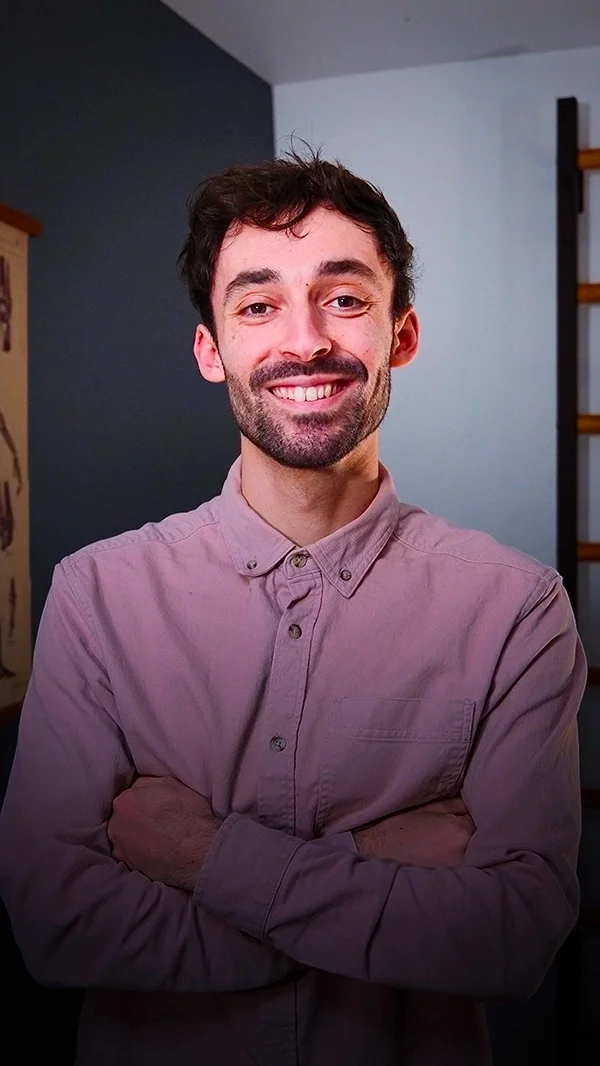 Nicolas Ménard, jeune homme souriant porte une chemise rose pâle, les bras croisés, dans un intérieur moderne avec un mur blanc et une étagère en bois en arrière-plan.