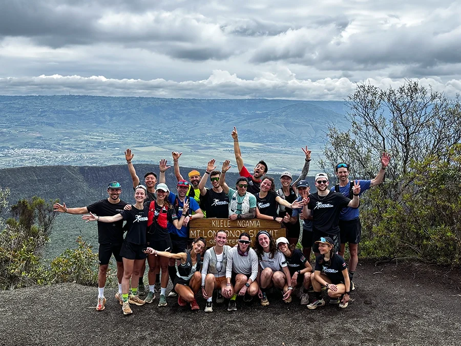 Groupe de coureurs souriantes et souriants lors d'une randonnée en montagne à Iten, avec un paysage vallonné en arrière-plan et un ciel nuageux.
