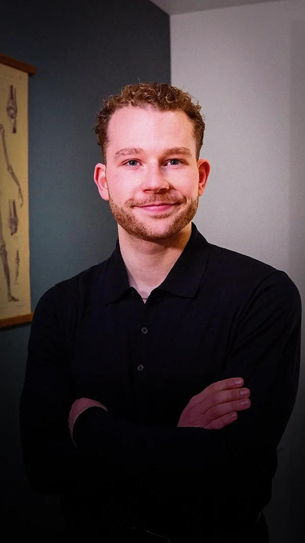 Louis Gronemberger, jeune homme souriant avec des cheveux courts et bouclés, portant une chemise noire, bras croisés, dans un intérieur avec une peinture en arrière-plan.