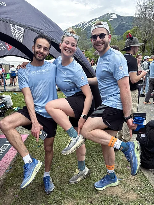Louis Gronemberger et 2 amis, portant des vêtements de trail, souriantes, se tenant sous une tente lors d'un événement en plein air avec des montagnes en arrière-plan. Ils montrent leurs nouvelles paires de Merrell.