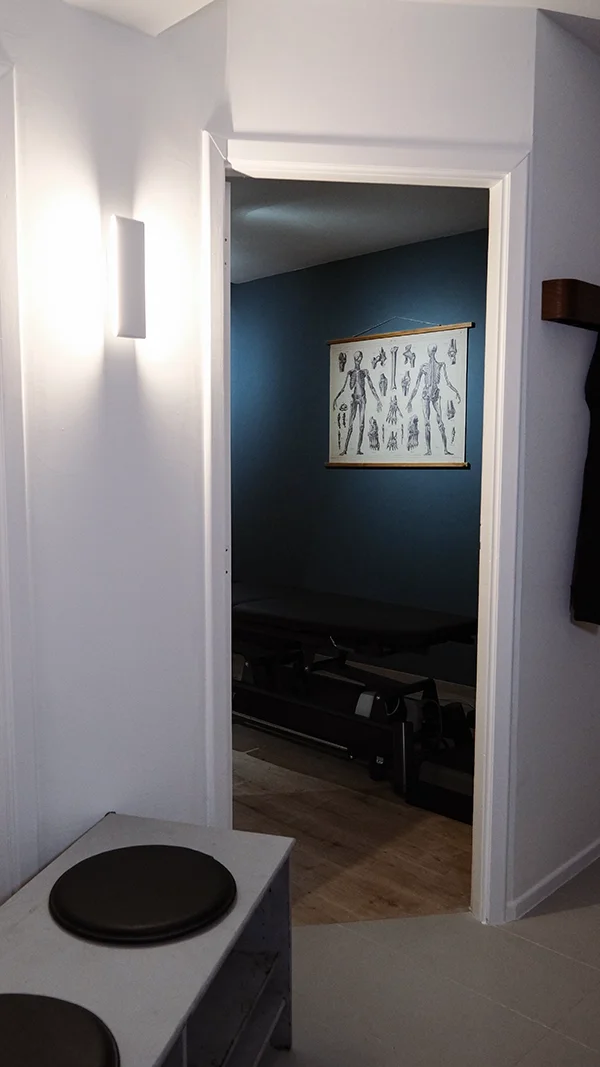Une pièce avec un mur bleu foncé, une affiche anatomique suspendue, un tapis de massage ou une table de traitement médical, et un éclairage mural, dans le cabinet BePerFit à Wezembeek-Oppem.