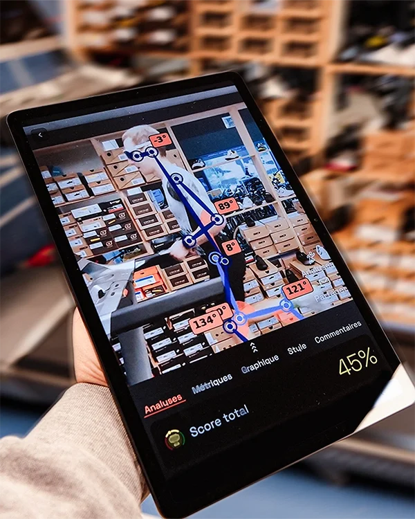 Une personne tient une tablette affichant une analyse d'image avec un fond de stock de chaussures en tiroirs. La tablette montre des angles et des mesures en degrés sur l'image. C'est une analyse de foulée avec Ochy chez Urban Tri Sports.
