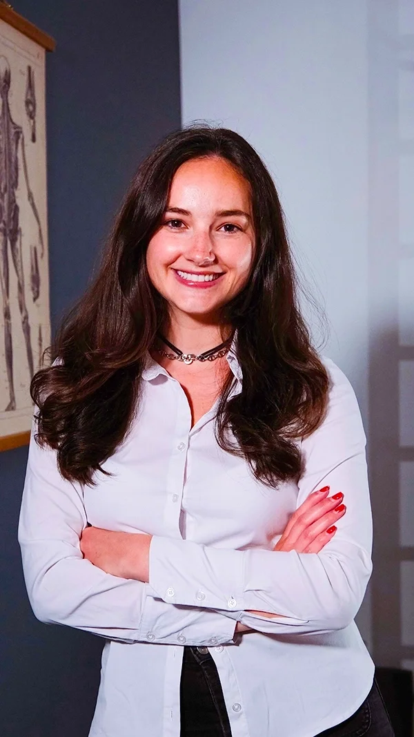 Marine Platiau, jeune femme souriante avec des cheveux longs et bruns, portant une blouse blanche, dans une salle avec un tableau anatomique au mur.
