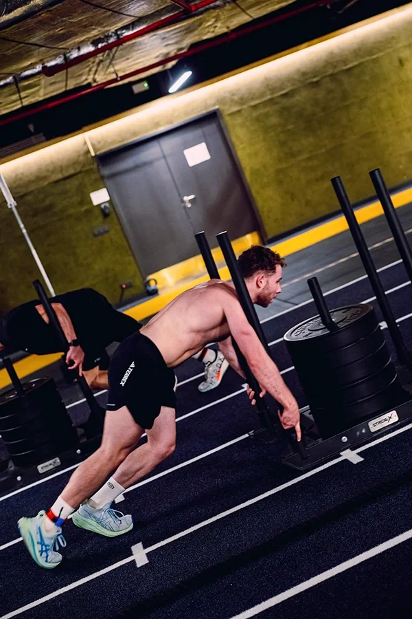 Un homme sportif en train de faire des exercices de musculation dans une salle de sport Hyrox, au Mix Brussels, avec des poids et un tapis de course autour.