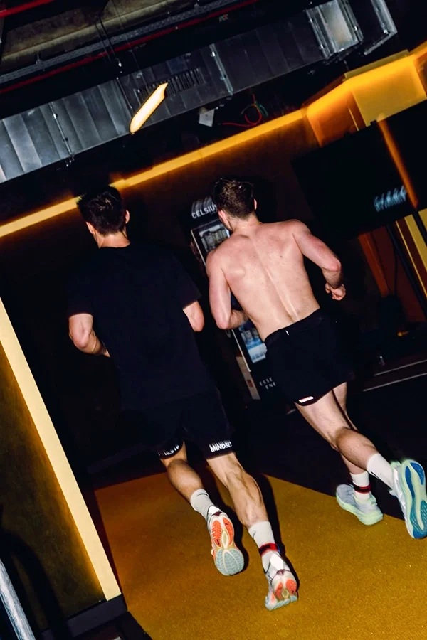 Deux hommes courent dans un bâtiment, l'un est torse nu et porte un short noir, l'autre porte un t-shirt noir et un short noir, tous deux portent des chaussures de sport. Ils s'entraînent pour de l'Hyrox au Mix Brussels.