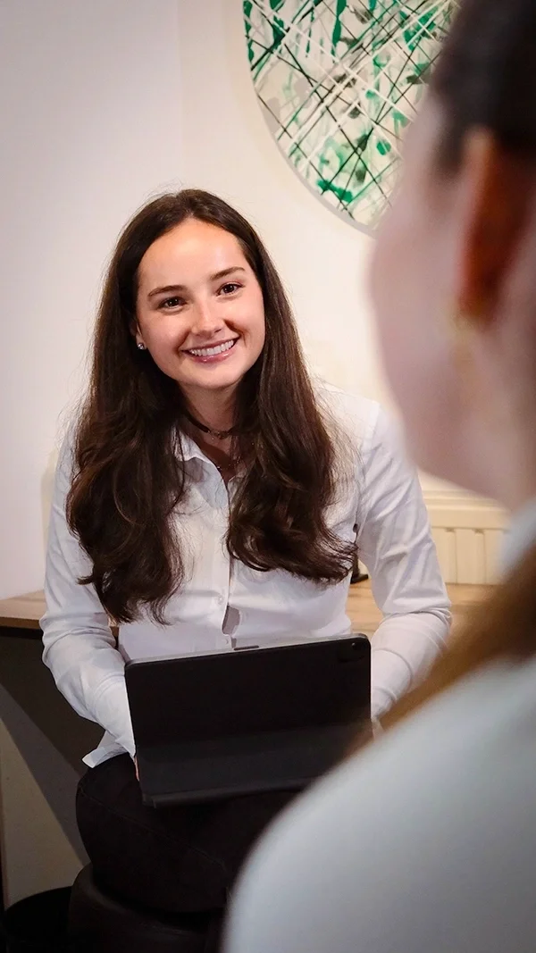 Une femme nutritionniste souriante avec des cheveux longs, portant une chemise blanche, lors d'une conversation avec une autre personne dans un intérieur.
