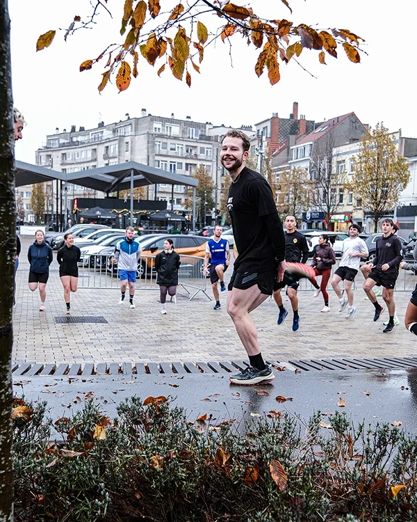 Un groupe de personnes en tenue de sport s'entraînent ou font du sport en extérieur dans une place urbaine, avec Louis Gronemberger en avant-plan sautant, sous un arbre avec des feuilles d'automne, lors d'un échauffement avec Onyx.