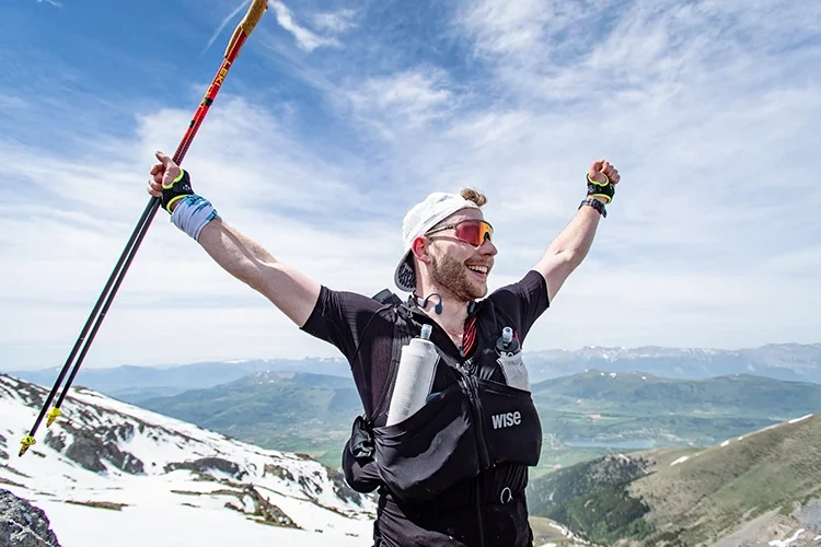 Louis Gronemberger, kiné et traileur souriant avec des lunettes de soleil, portant un sac à dos, en haut d'une montagne enneigée, célébrant avec les bras levés, en arrière-plan un ciel bleu et des montagnes.