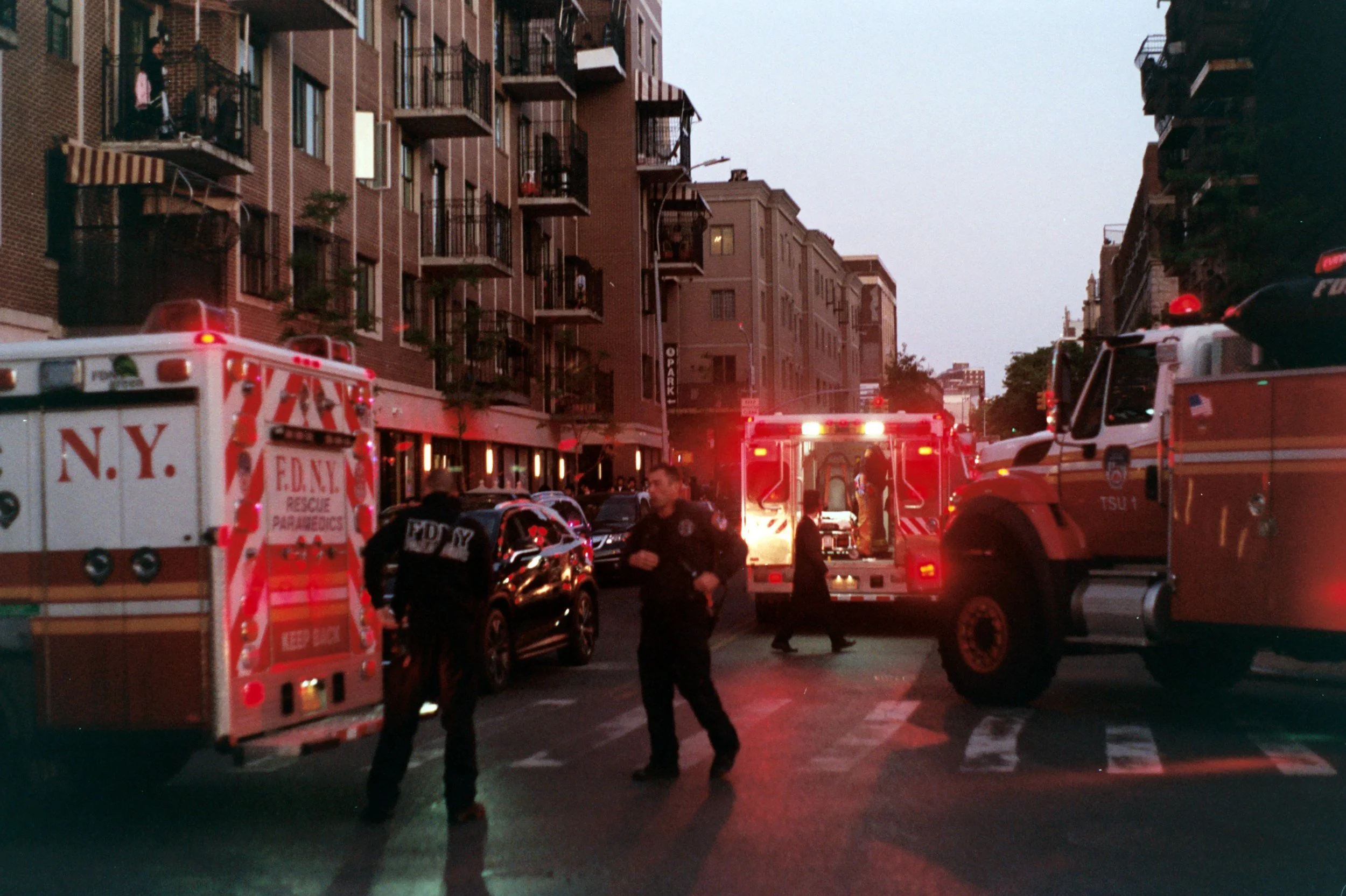 Emergency response scene on a city street with fire trucks, police officers, and rescue personnel attending to an incident amid residential buildings at dusk.