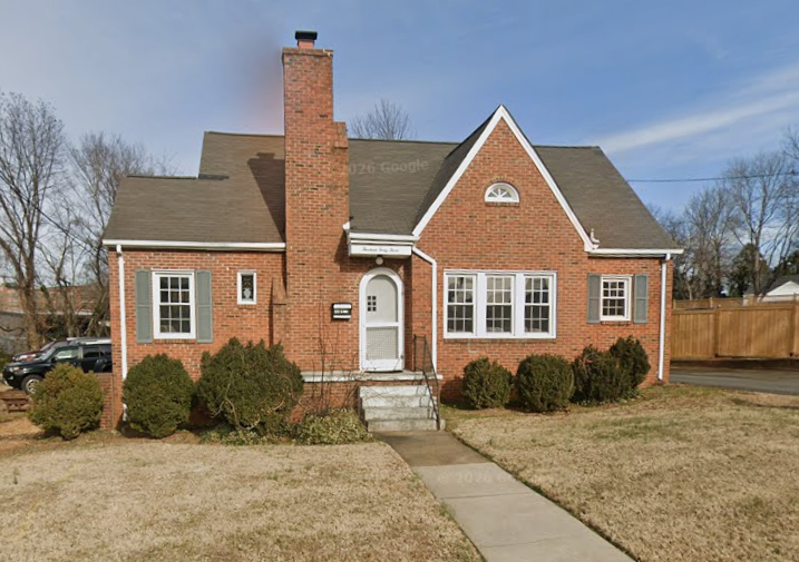 A brick house with a gabled roof, front steps, and a chimney, surrounded by a small lawn and bushes, with a wooden fence on the right and a car parked in the background.