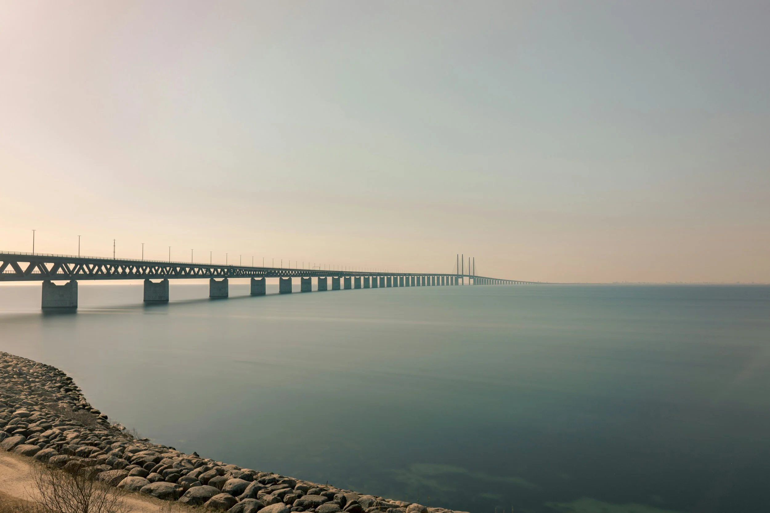 A long bridge extends over calm water with a rocky shoreline in the foreground and a clear sky.
