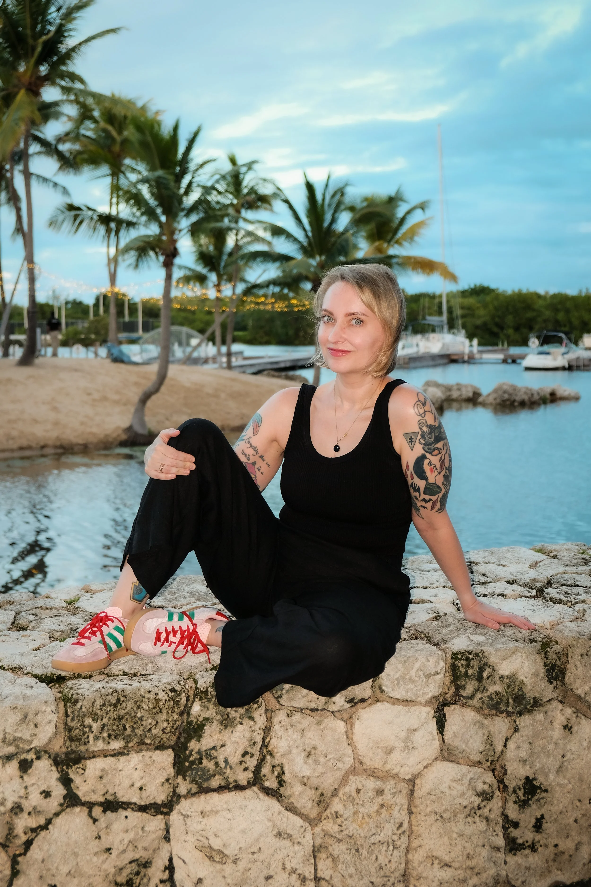 A woman with tattoos sitting on a stone wall near water, with palm trees and boats in the background, during dusk.