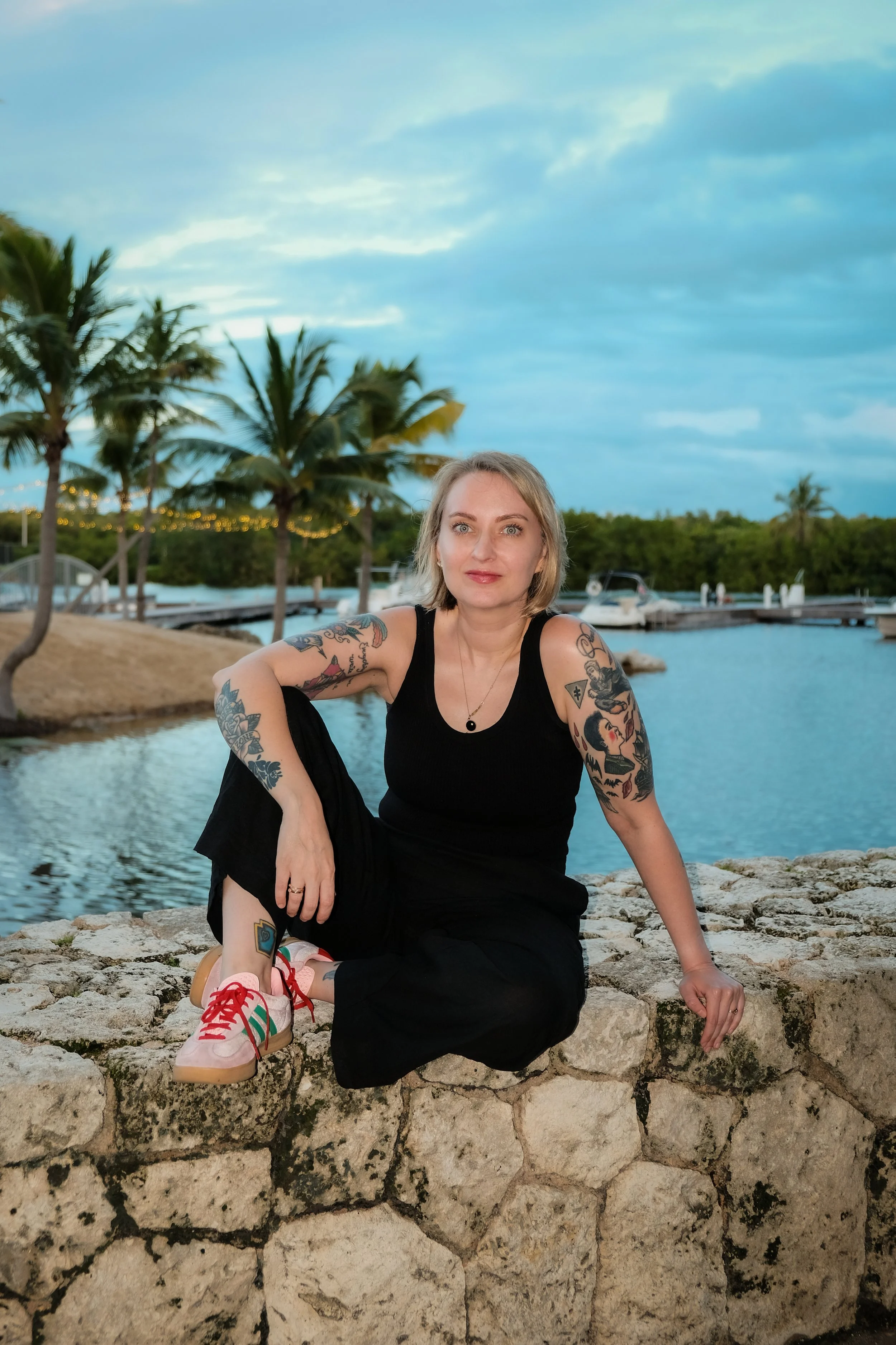 A woman with tattoos on her arms and short blonde hair, dressed in black, sitting on a stone wall near a body of water with palm trees and boats in the background during dusk.