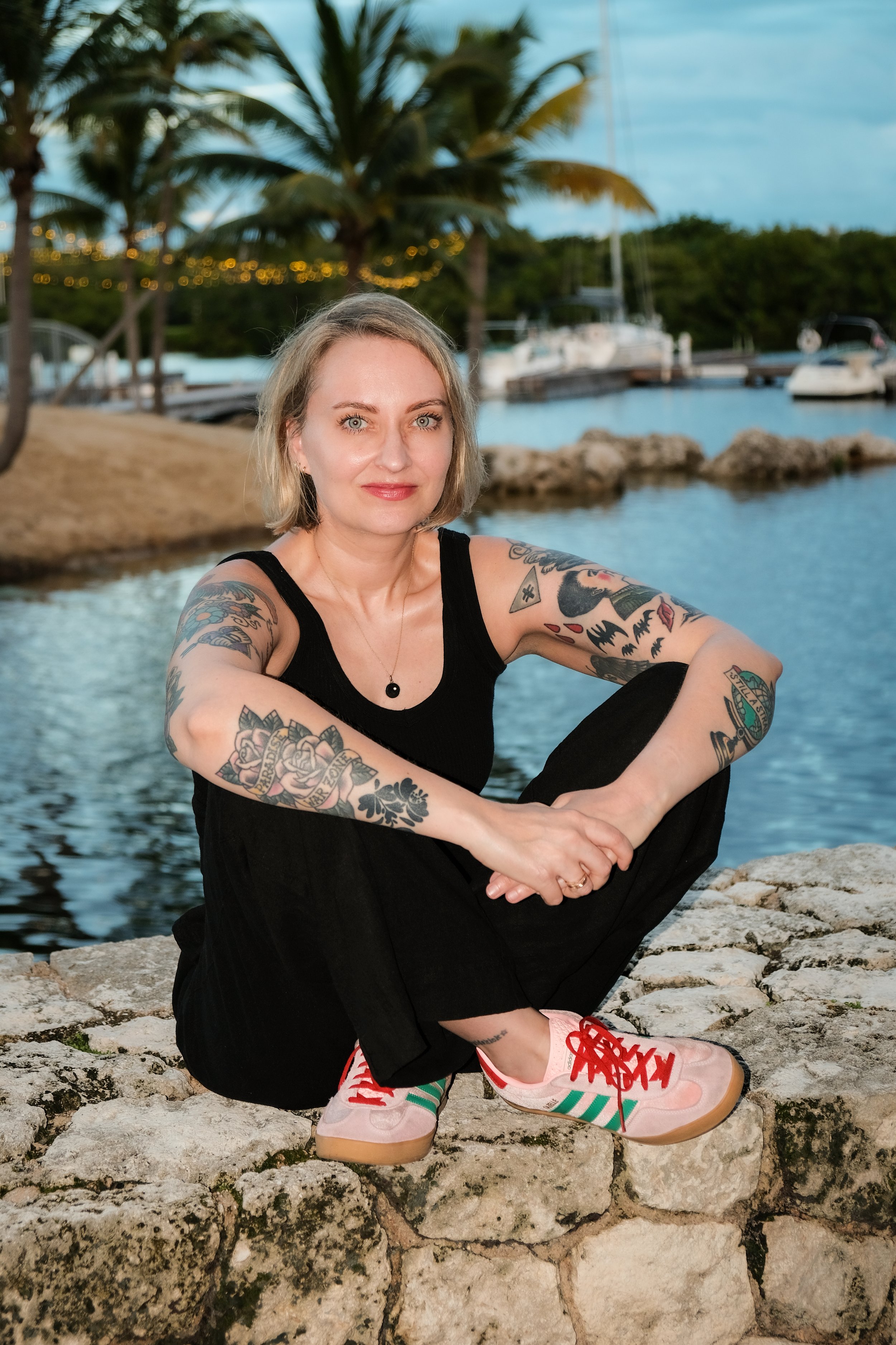 A woman with tattoos on her arms sitting on a stone wall by a marina with sailboats and palm trees in the background.