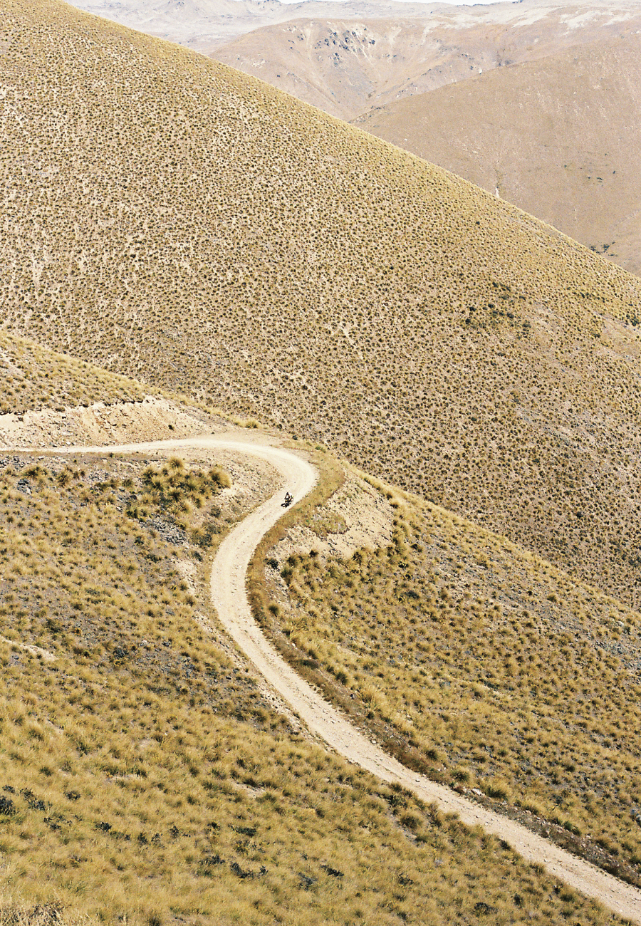 A winding dirt road through a dry, mountainous landscape with sparse vegetation and rolling hills in the distance.