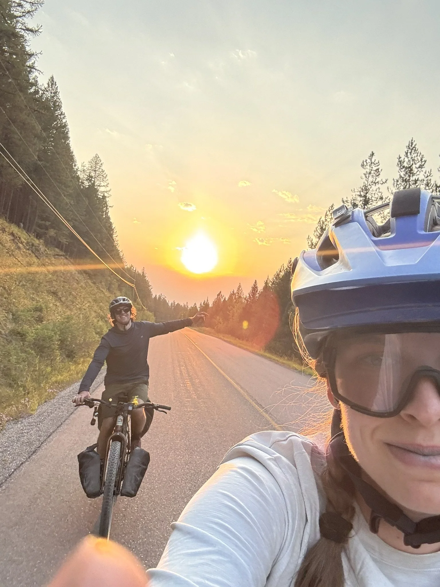 Two women wearing bicycle helmets and sunglasses taking a selfie during a sunset ride on a rural road surrounded by trees.