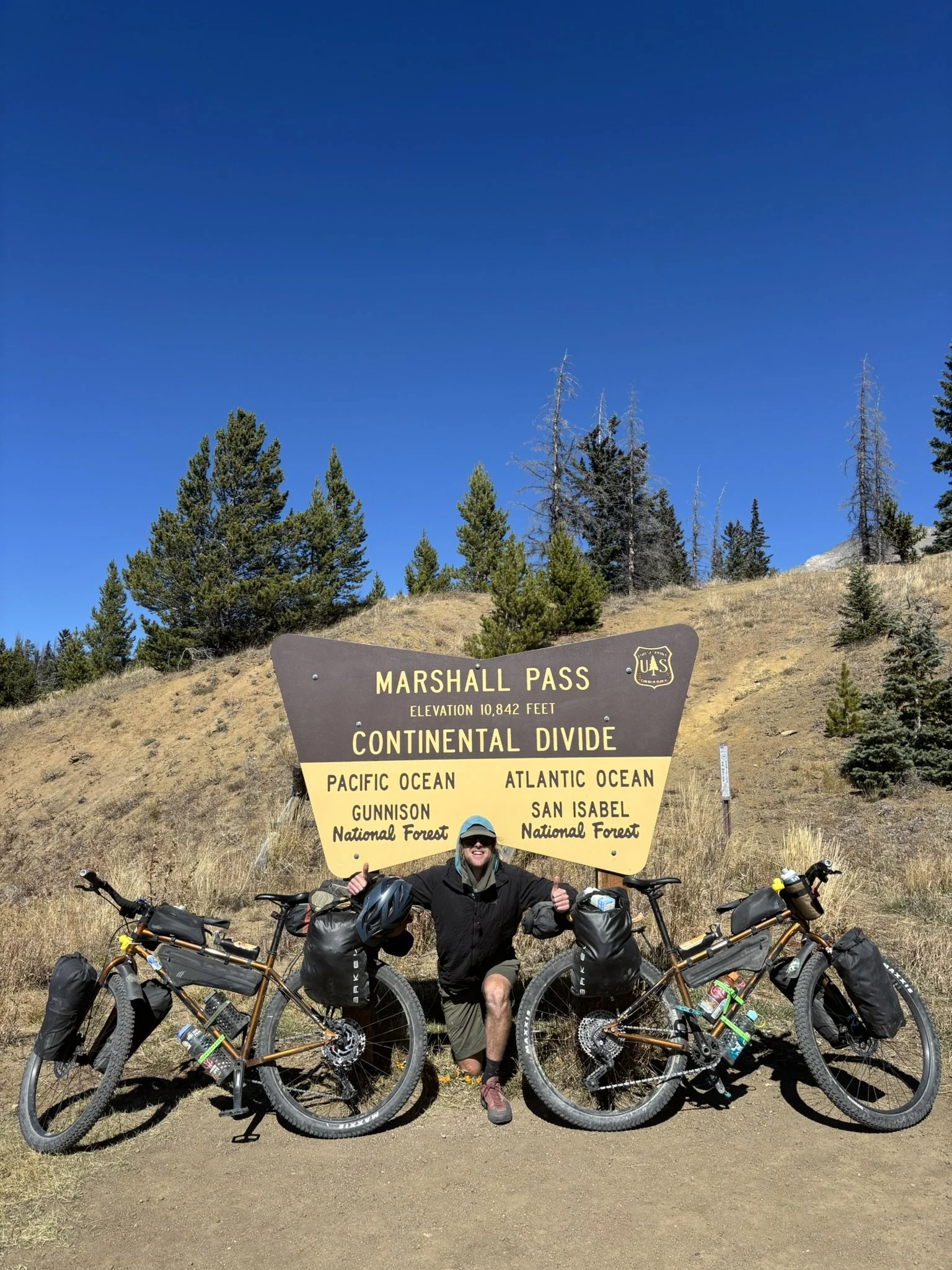 Person kneeling in front of a sign at Marshall Pass with two loaded bicycles on either side, under a blue sky and trees.