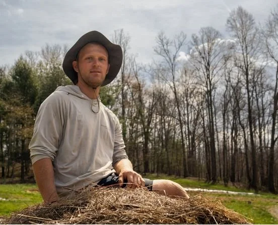 A man in a wide-brimmed hat and light-colored hoodie sitting outdoors near a pile of sticks or branches, with a background of leafless trees and a cloudy sky.