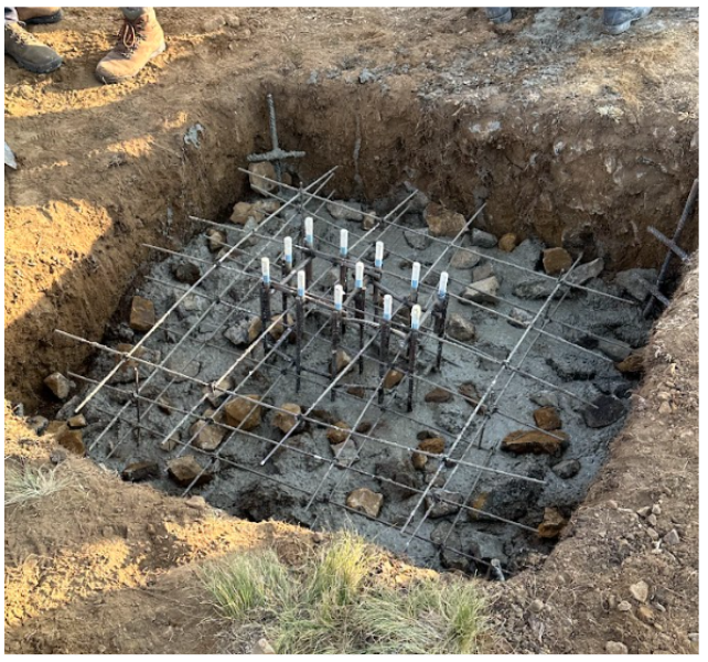 A construction site showing a foundation with rebar and concrete, surrounded by excavated dirt.
