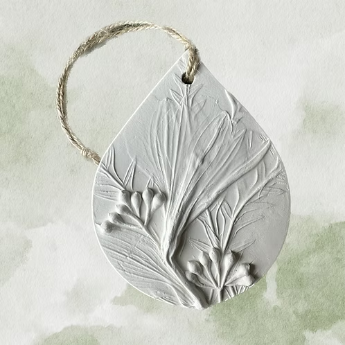 A round, white, ceramic ornament with a raised floral design, attached to a piece of twine. Botanical Bas Relief Essential Oil Diffuser.