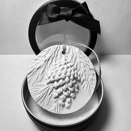 A white pinecone Christmas ornament with detailed textured pinecone and leaf design, placed in an open black round box with a transparent lid and black ribbon bow.