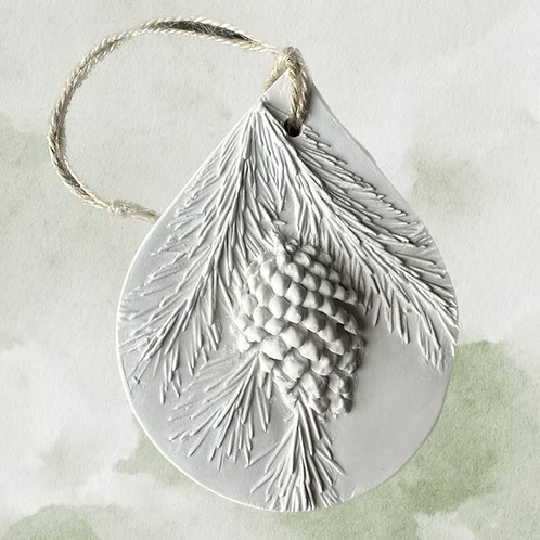 White ceramic ornament with pine cone and pine needle carvings, attached to a looped string for hanging. Botanical Bas Relief Essential Oil Diffuser.