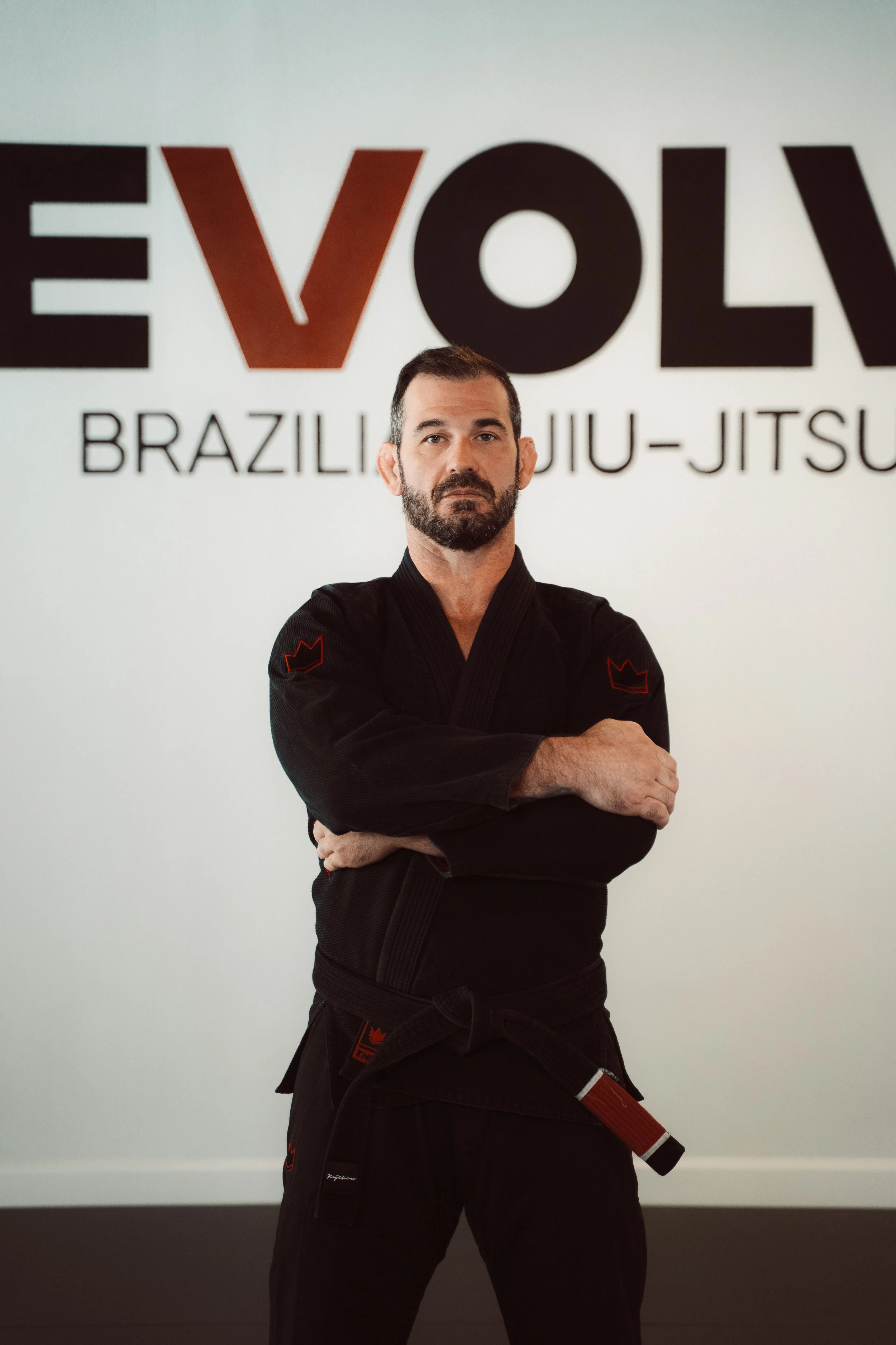 A man in a black Brazilian Jiu-Jitsu gi standing with arms crossed in front of a wall with the words 'EVOLV Brazilian Jiu-Jitsu'.