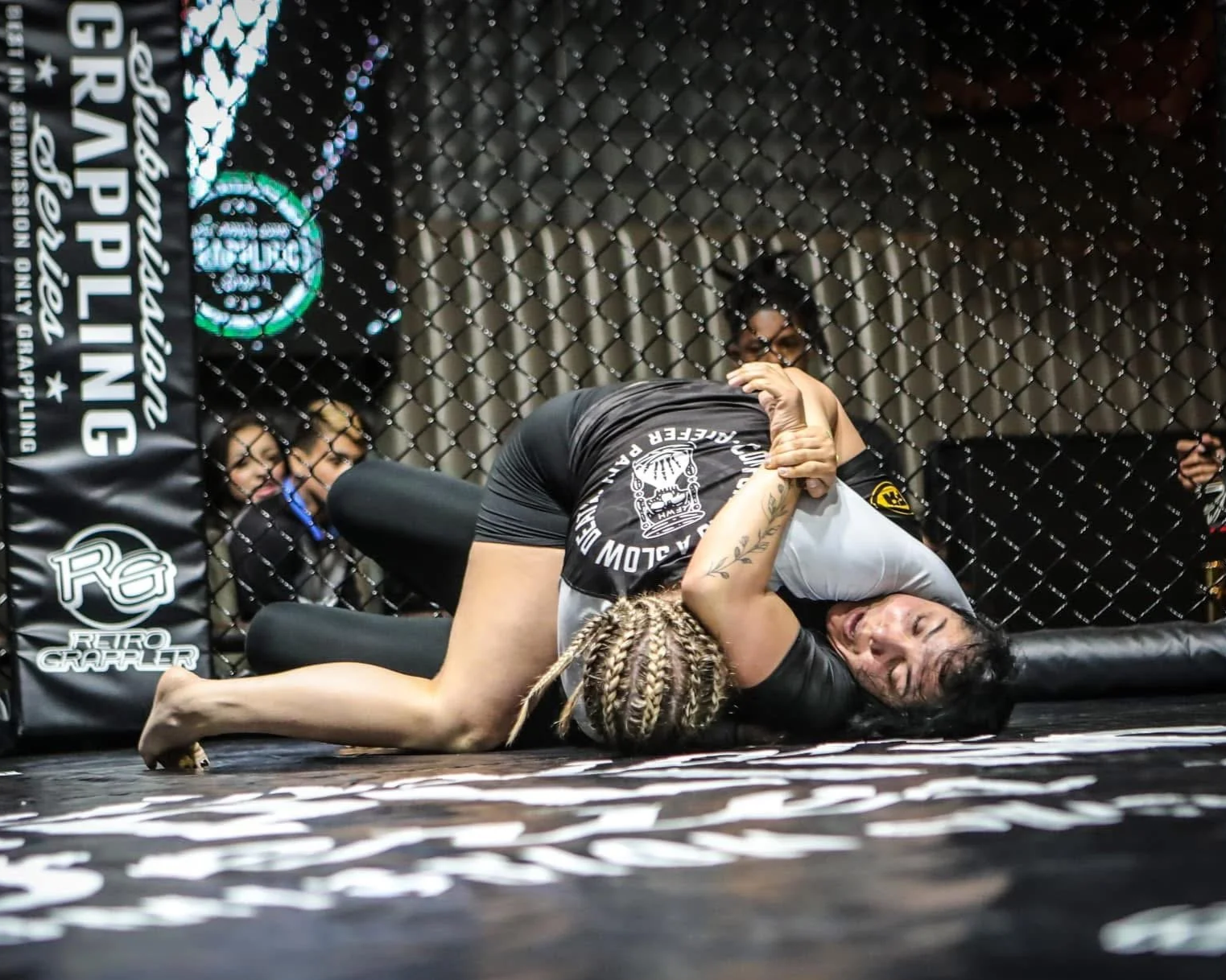 Two women grappling inside a mixed martial arts cage, with one woman in mount and the other trying to escape, while several spectators watch in the background.