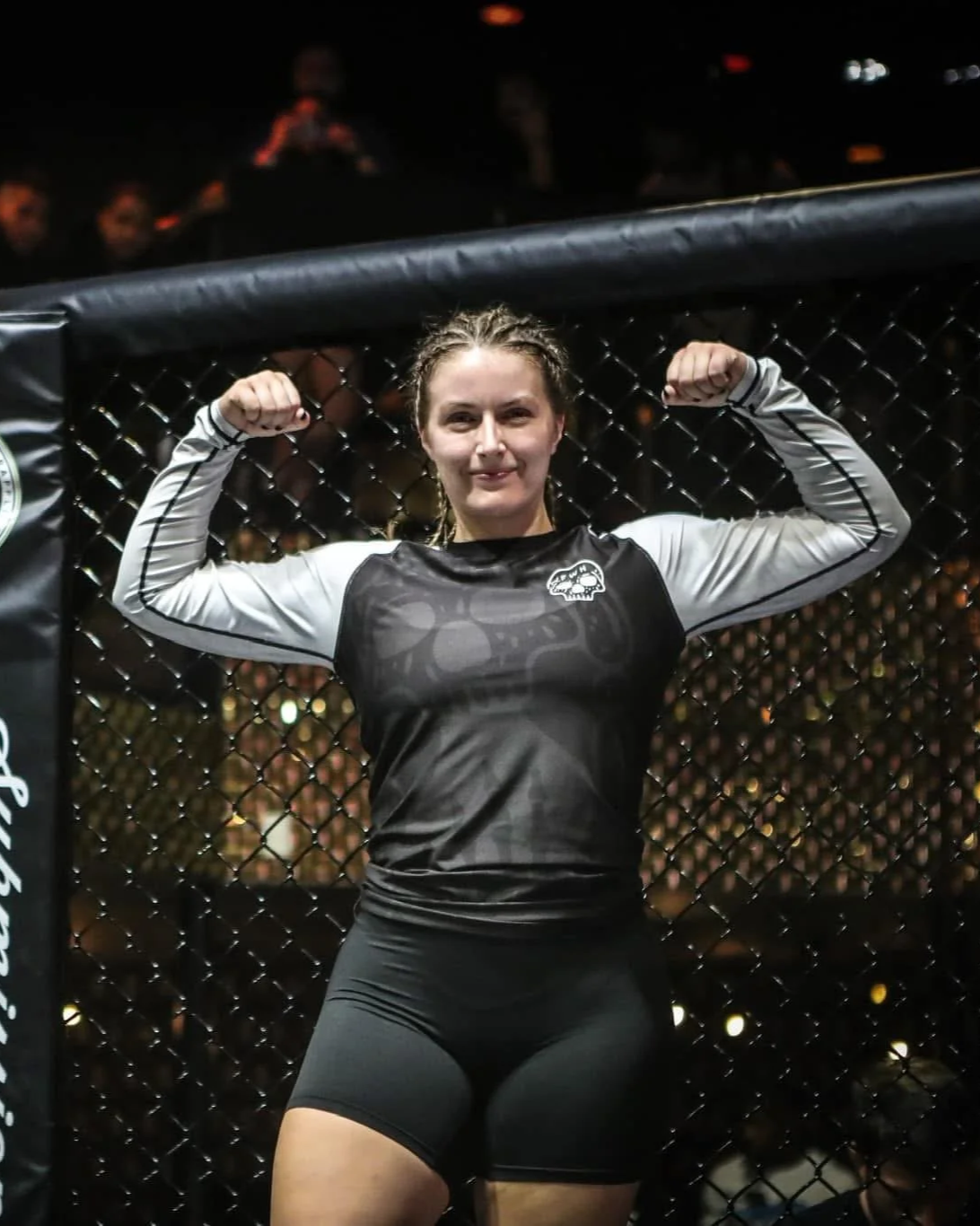 Female mixed martial artist inside a fighting cage, flexing her biceps and smiling