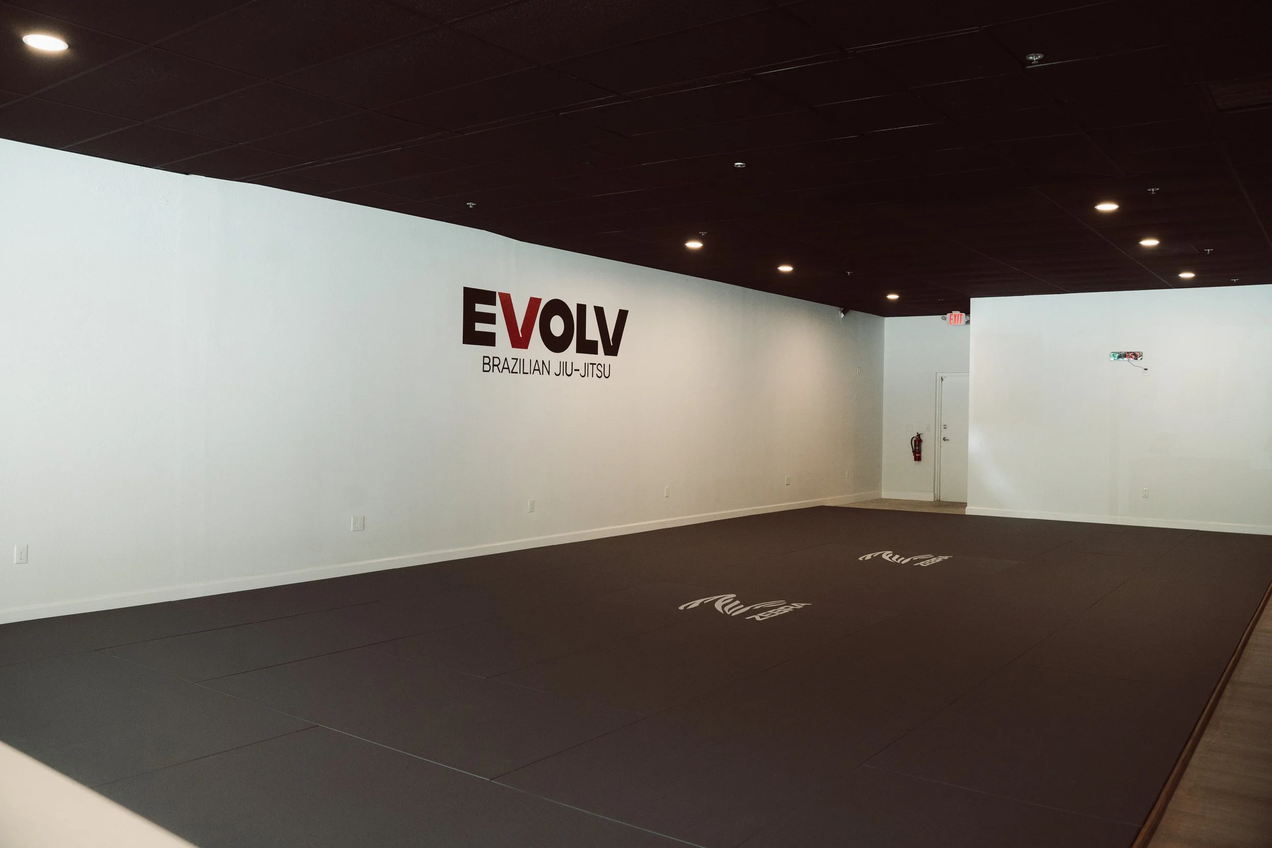 Empty Brazilian Jiu-Jitsu training mat room with EVOLVE logo on the wall, dim lighting, and a fire extinguisher near the door.