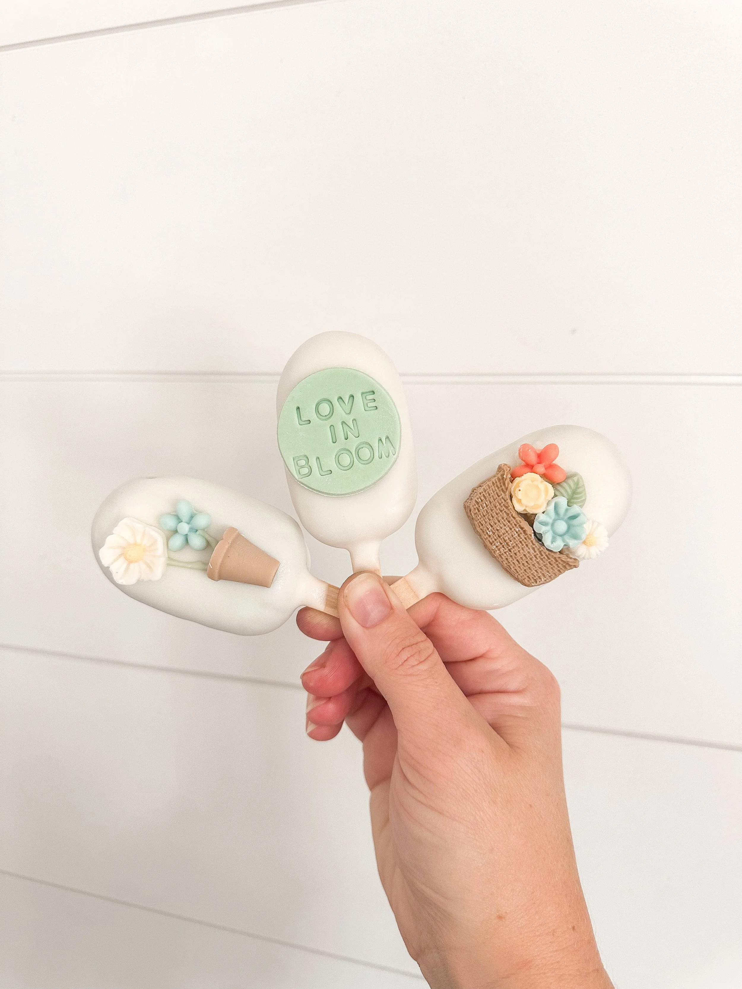 Hand holding three decorated cake pops with flowers, a small pot, and a basket on top of each, against a white background.