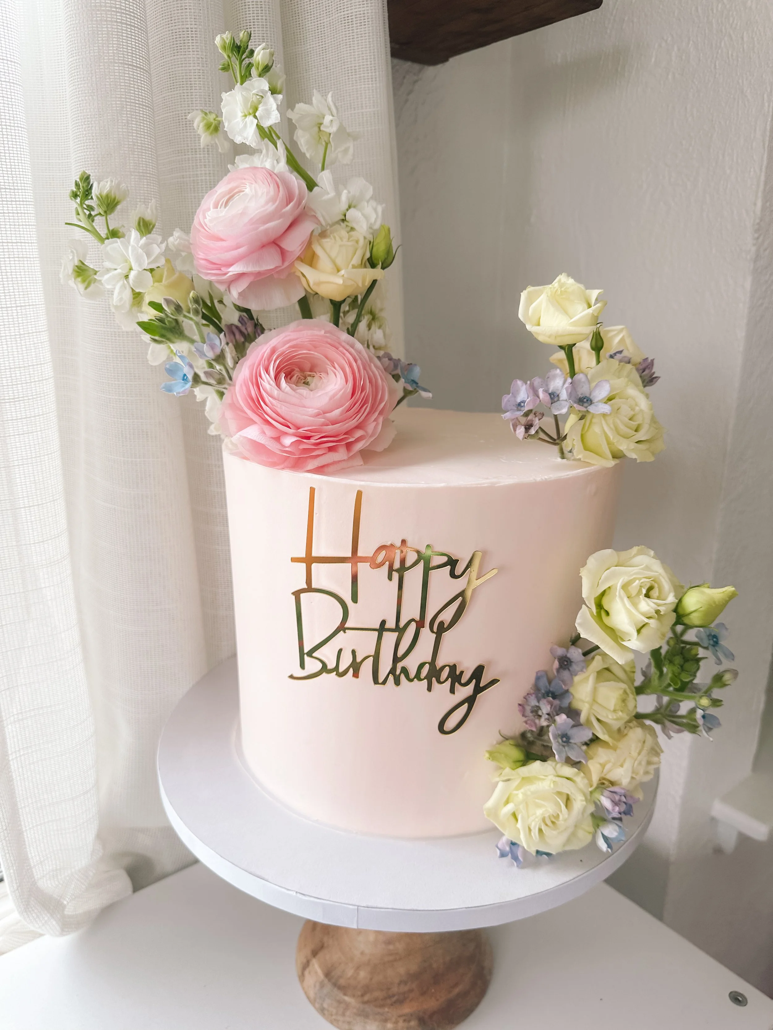 A pink birthday cake decorated with fresh white, pink, and purple flowers and a gold 'Happy Birthday' topper, placed on a white stand.