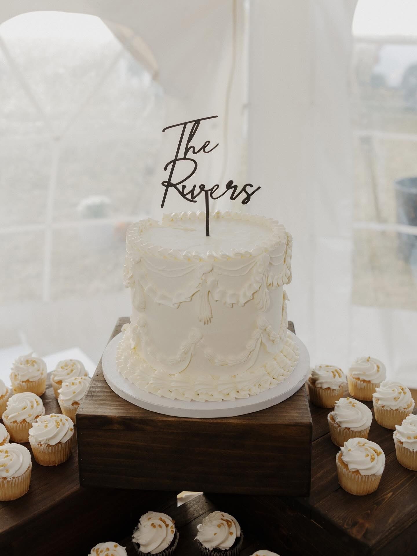 A white frosted cake with decorative piping is displayed on a wooden stand, with cupcakes surrounding it. The cake features a black topper that spells 'The Ruppers' in cursive. The scene appears to be set at an event or celebration.