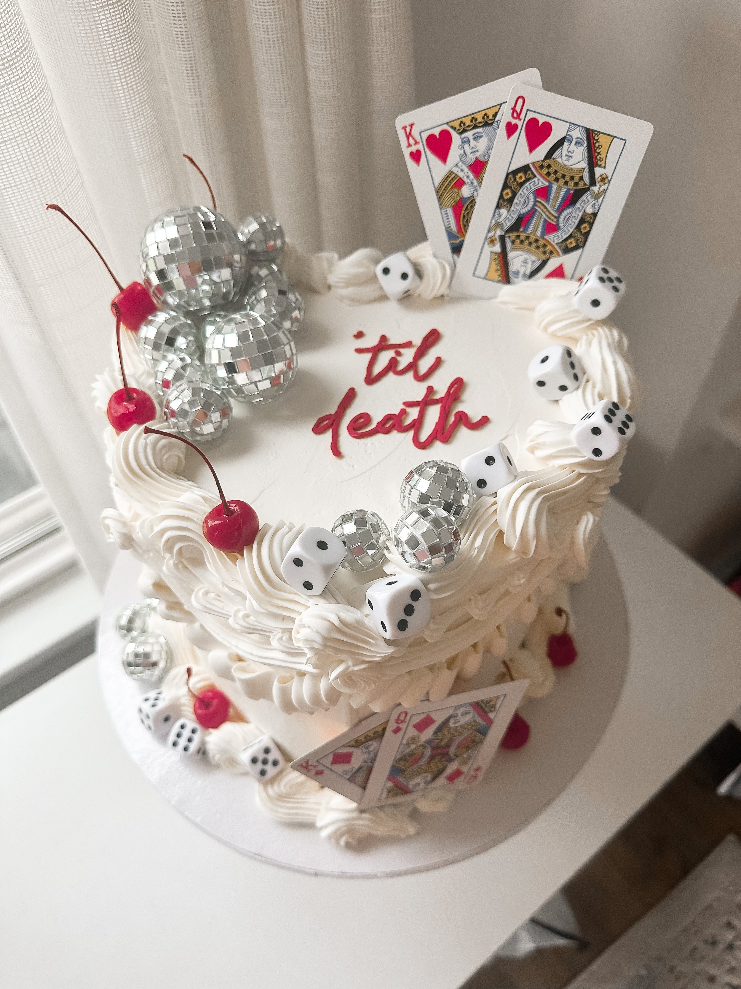 Celebration cake decorated with playing cards, cherries, dice, and disco balls with the words 'til death' written on top.