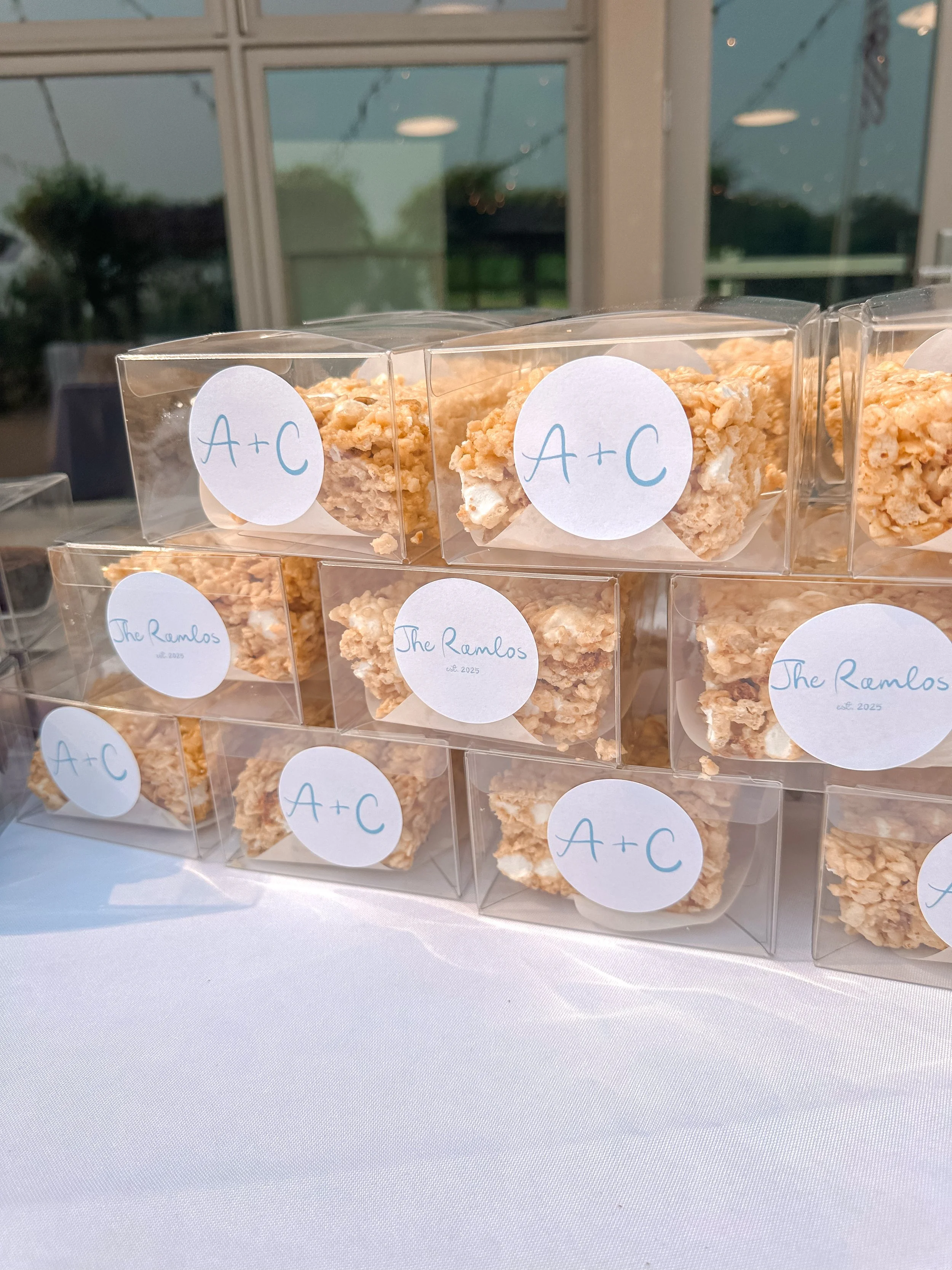 Stacked clear favor boxes with oatmeal bars labeled 'A+C' and 'The Ramlos' on a table at a reception or event.