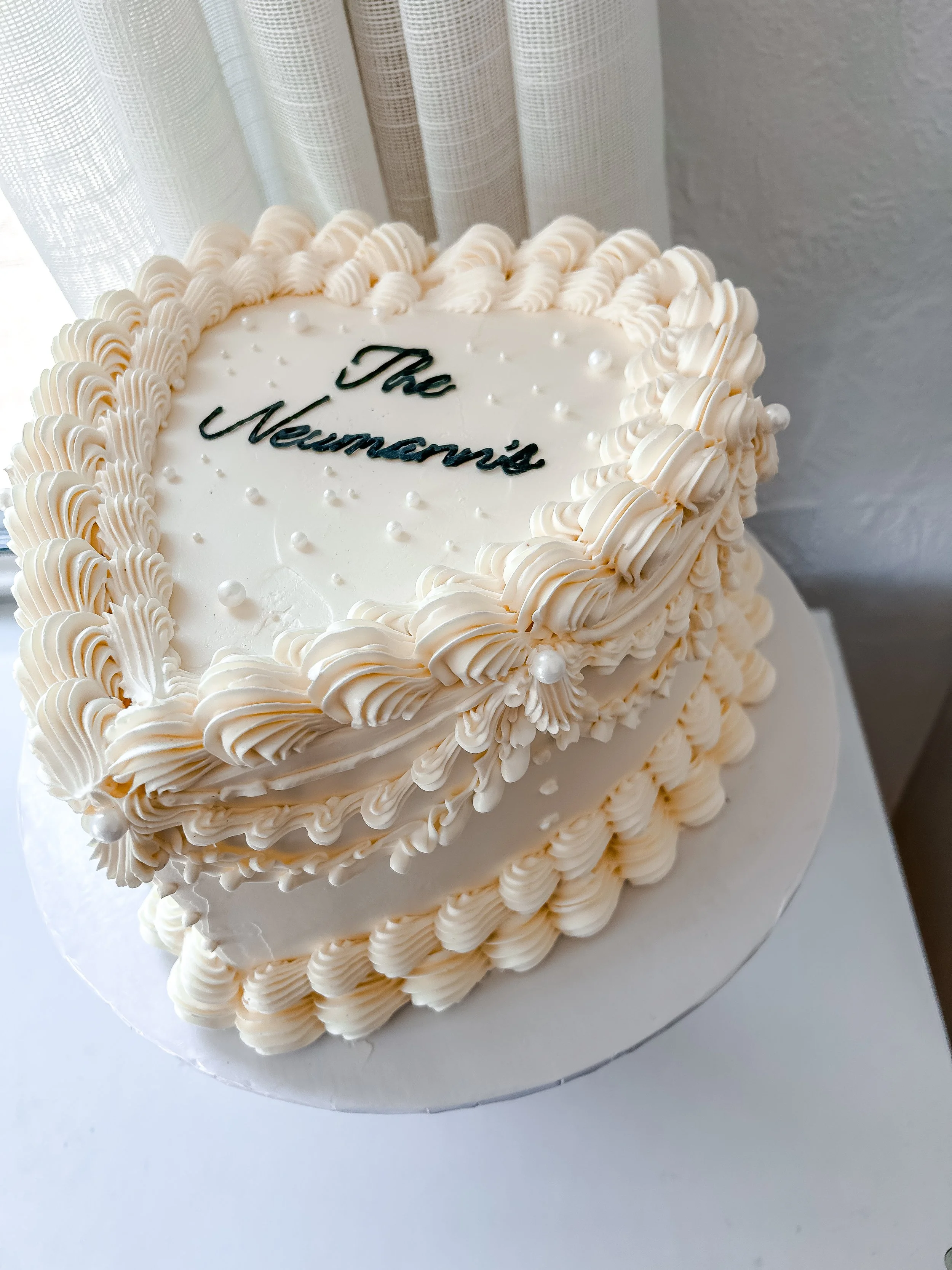 Cream-colored heart-shaped cake decorated with piped frosting, pearls, and a message reading 'The Neumann's' on top.