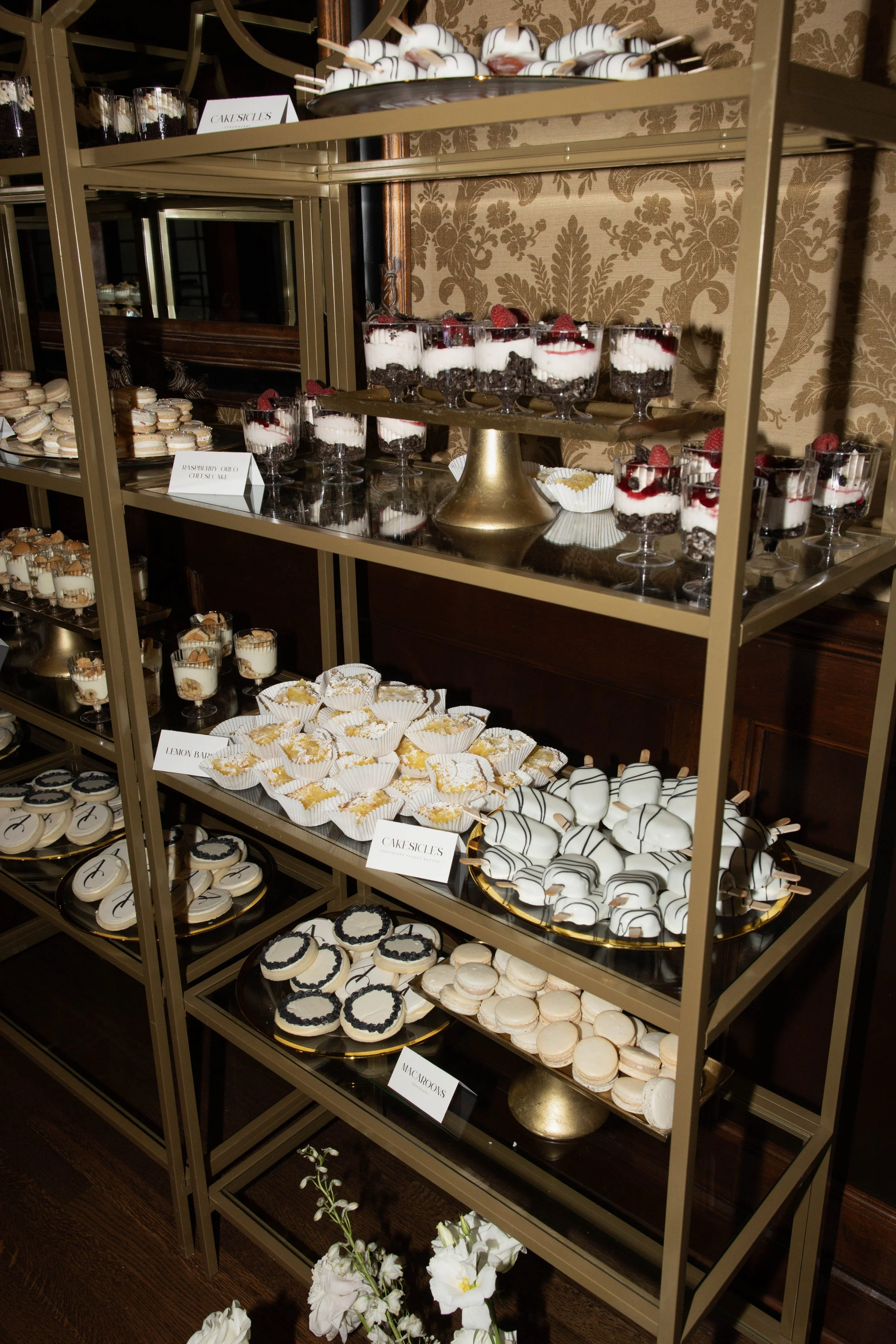 Display of various desserts including mini cakes, parfaits, macarons, and decorated cookies on a multi-tiered gold and glass dessert stand at a buffet. Signs indicate flavors like 'Cakesicles,' 'Lemon Bar,' and 'Macarons'.