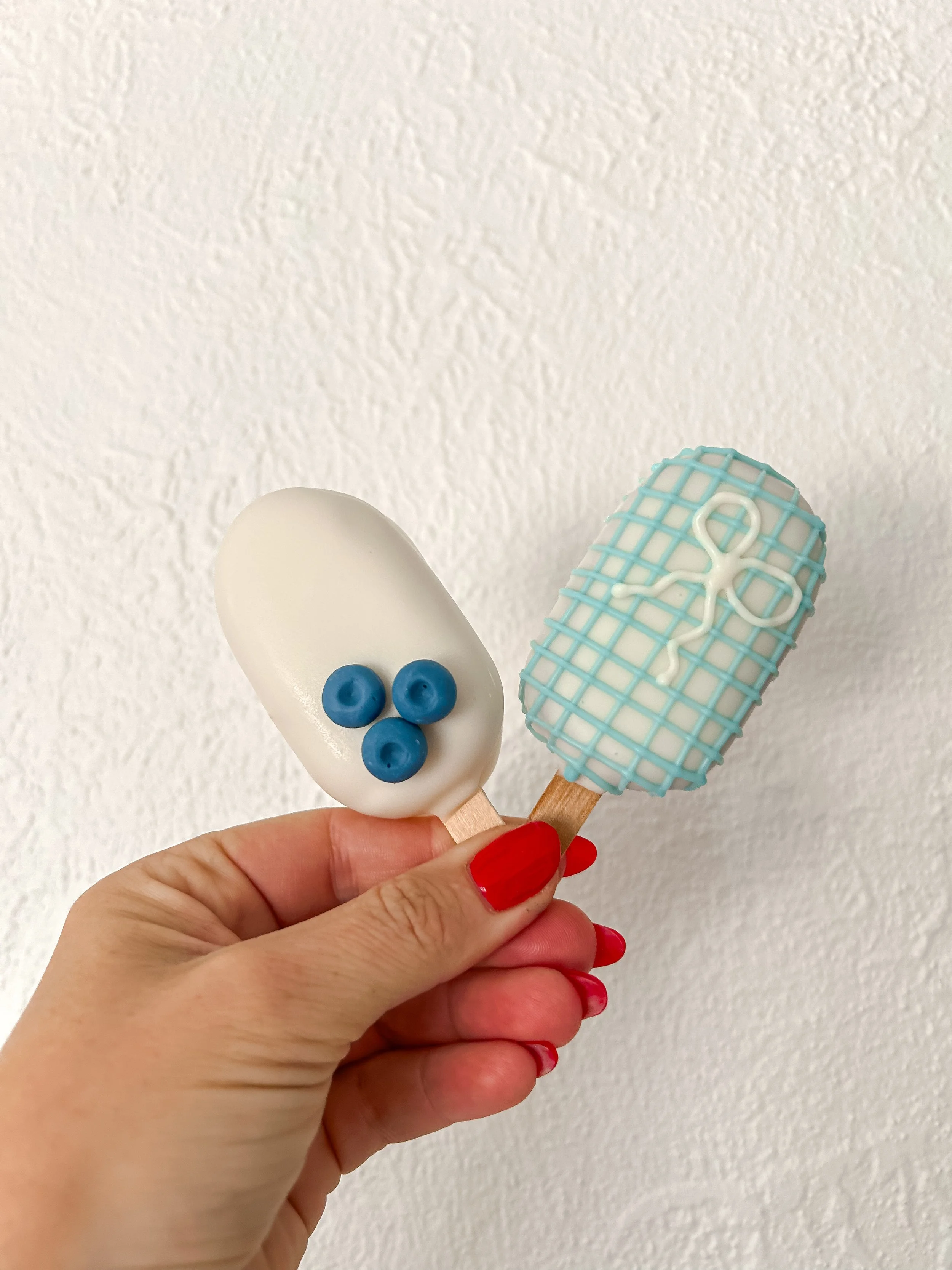 Two ice cream bars with decorative toppings held by a hand with red nail polish against a white textured wall.