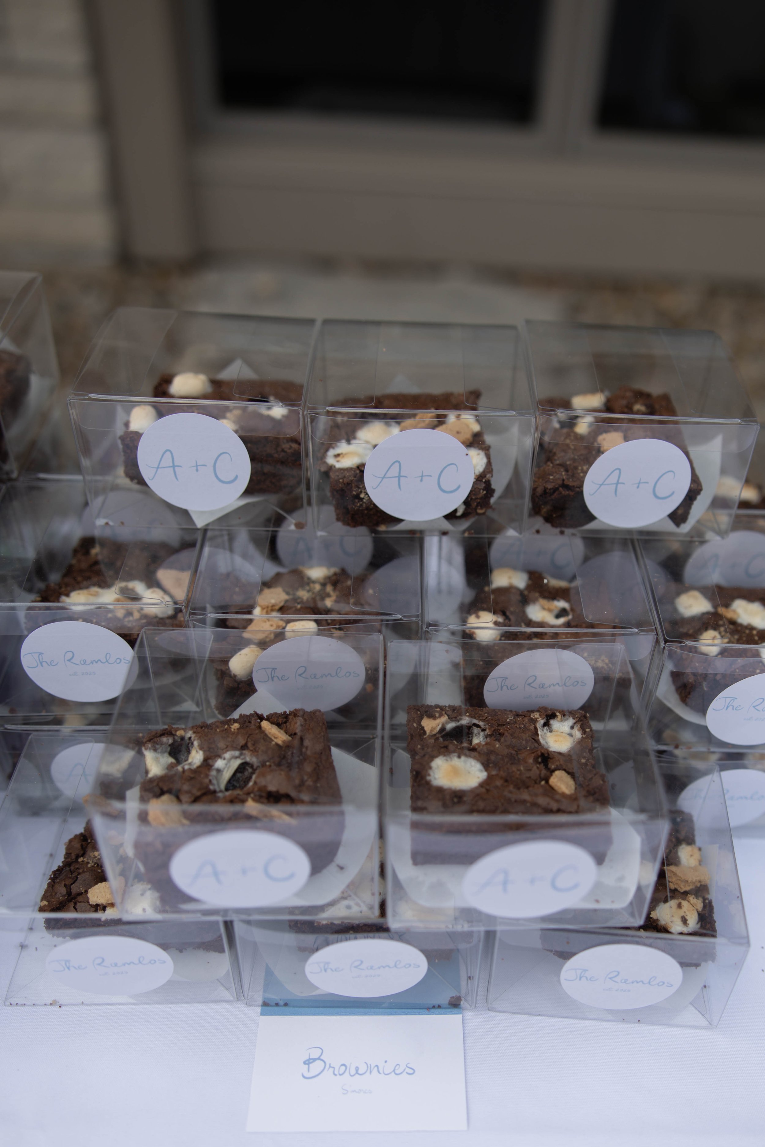 Several individually packaged brownie squares in clear plastic boxes on a white surface, labeled with white stickers reading 'A+C' and 'The Ramlos'. A handwritten sign below reads 'Brownies'.